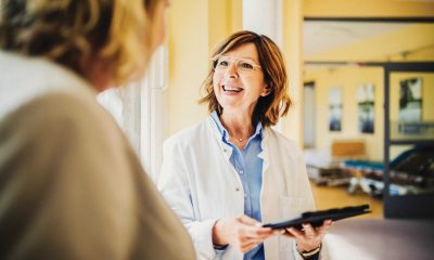 Female patient facing away from the camera talking to a  female healthcare provider in a lab coat holding a clipboard. 