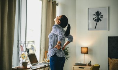 Adult woman stands in front of a desk, stretching her back.
