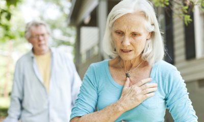 Older woman facing the camera puts her hand to her chest in sudden pain with older man in background looking concerned