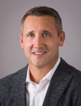 Photo of Garrett OBrien in front of a gray background. He is wearing a white button up shirt and dark gray suit jacket. 