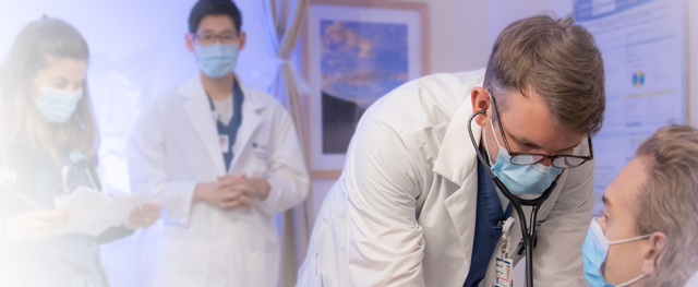 A resident examines a patient in a clinical setting while other providers take notes in the background
